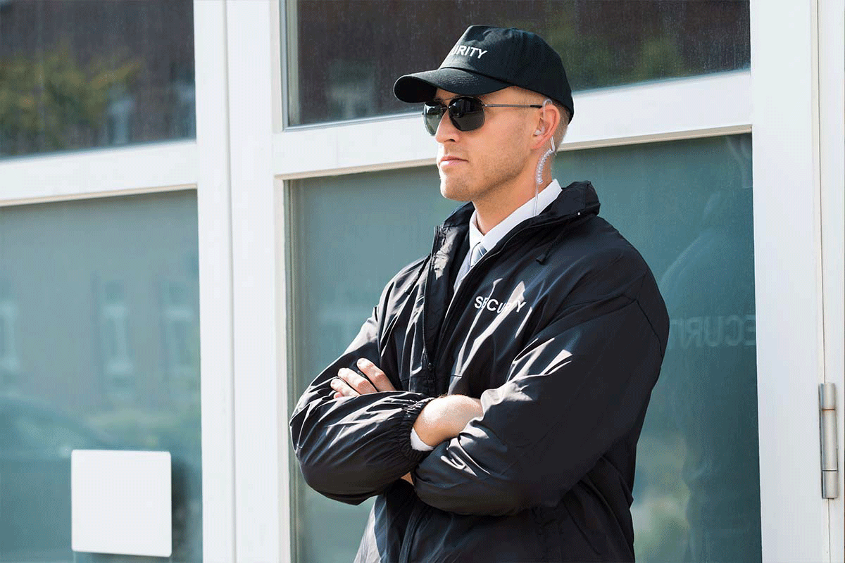 Security guard in uniform with crossed arms, wearing sunglasses and an earpiece, representing professional security services on duty.